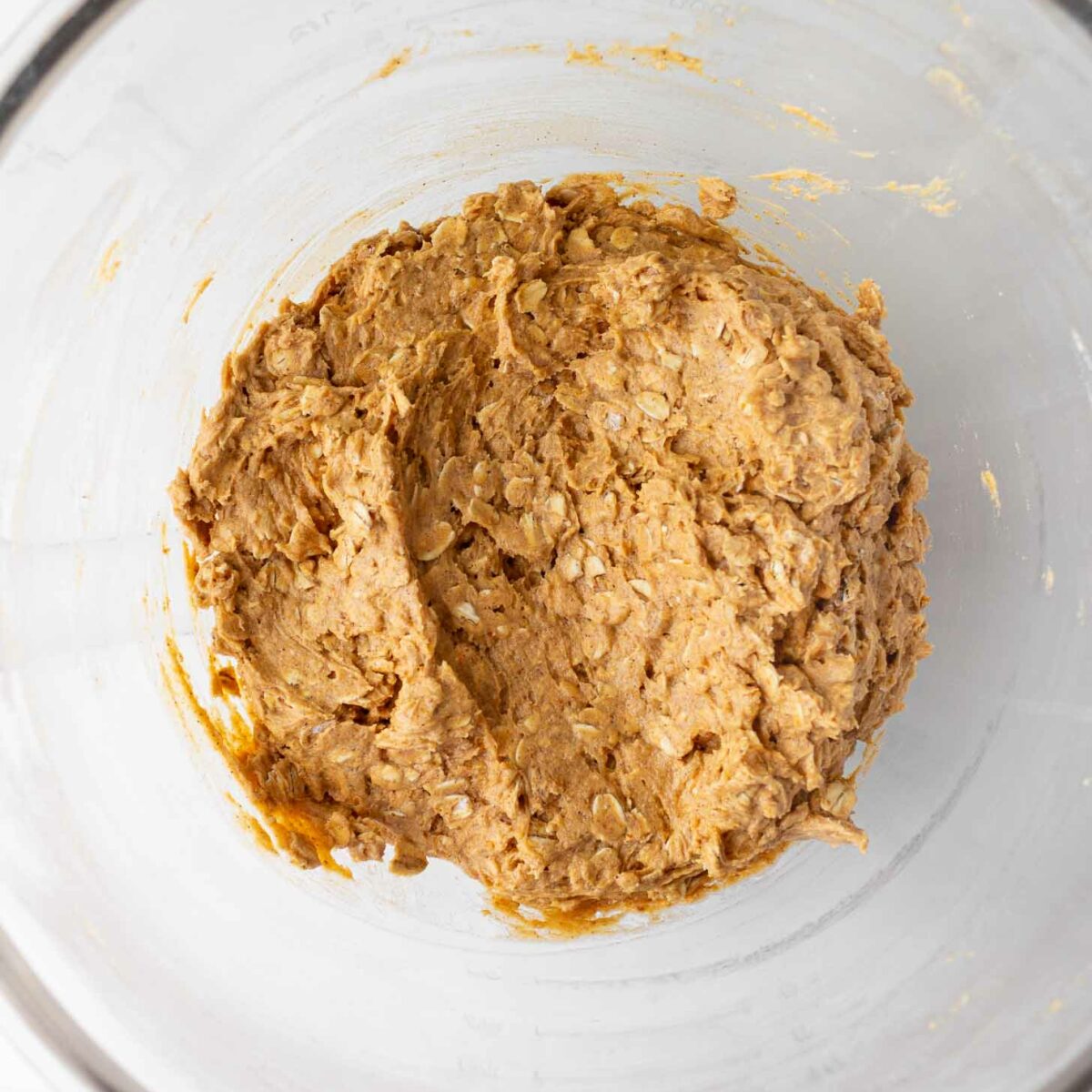A large glass bowl containing a thick, light brown oatmeal cookie dough mixture, viewed from above on a white surface.