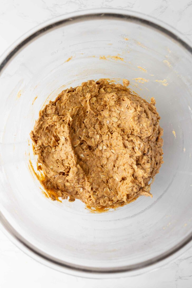 A large glass bowl containing a thick, light brown oatmeal cookie dough mixture, viewed from above on a white surface.