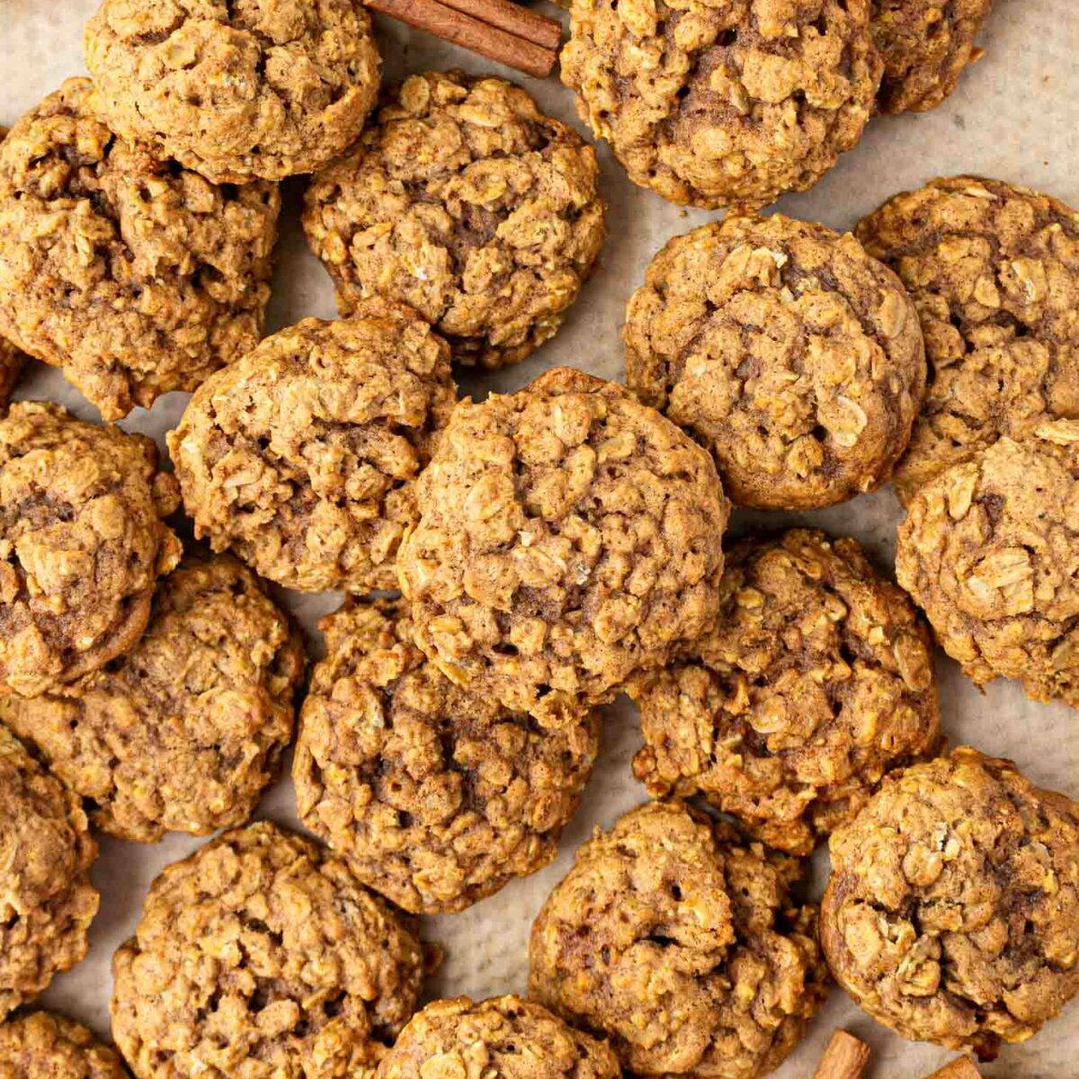 A batch of pumpkin oatmeal cookies is scattered on parchment paper with a few cinnamon sticks placed among them. The cookies appear soft and have a rustic, homemade texture.