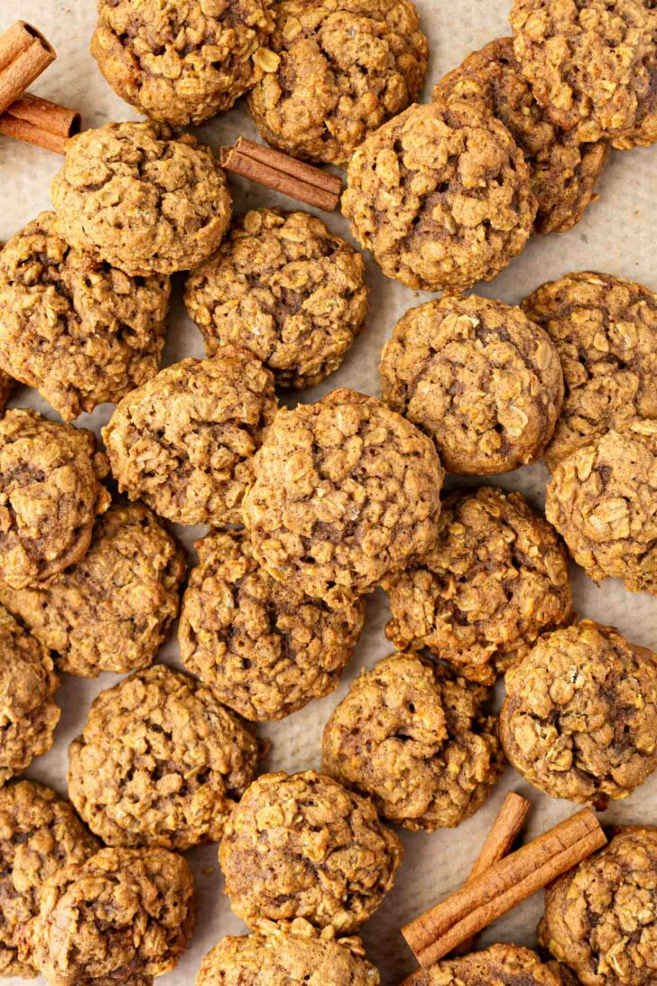 A batch of pumpkin oatmeal cookies is scattered on parchment paper with a few cinnamon sticks placed among them. The cookies appear soft and have a rustic, homemade texture.