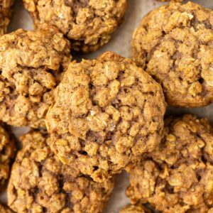 Close-up view of several pumpkin oatmeal cookies with a golden-brown color and a slightly rough, chunky texture, stacked closely together on a parchment-lined surface.