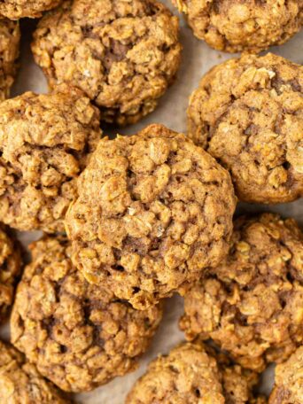 Close-up view of several pumpkin oatmeal cookies with a golden-brown color and a slightly rough, chunky texture, stacked closely together on a parchment-lined surface.