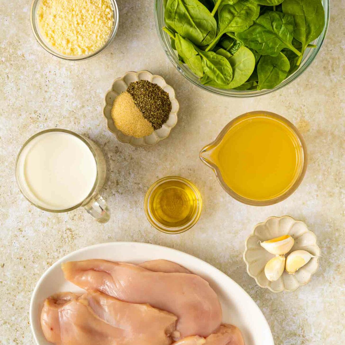 Top-down view of raw chicken breasts on a plate, fresh spinach in a bowl, grated cheese, cream, olive oil, chicken broth, spices, and garlic cloves arranged on a light surface.