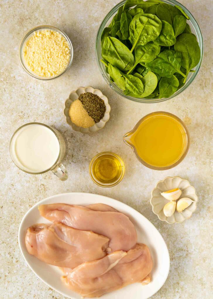 Top-down view of raw chicken breasts on a plate, fresh spinach in a bowl, grated cheese, cream, olive oil, chicken broth, spices, and garlic cloves arranged on a light surface.