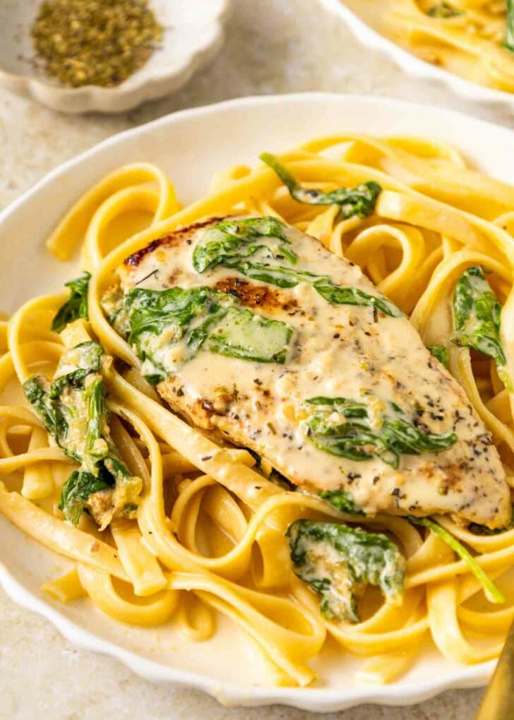A plate of fettuccine pasta topped with a creamy chicken breast, spinach, and a rich sauce, garnished with herbs. A small dish of seasoning sits in the background.