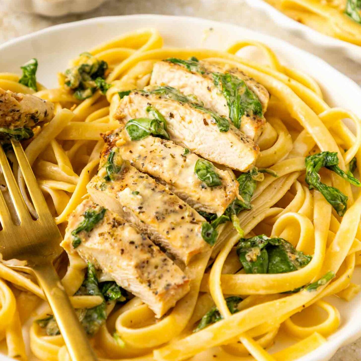 A plate of fettuccine pasta topped with sliced seasoned chicken breast and wilted spinach, served with a gold fork. A small dish of herbs and another plate of pasta are in the background.