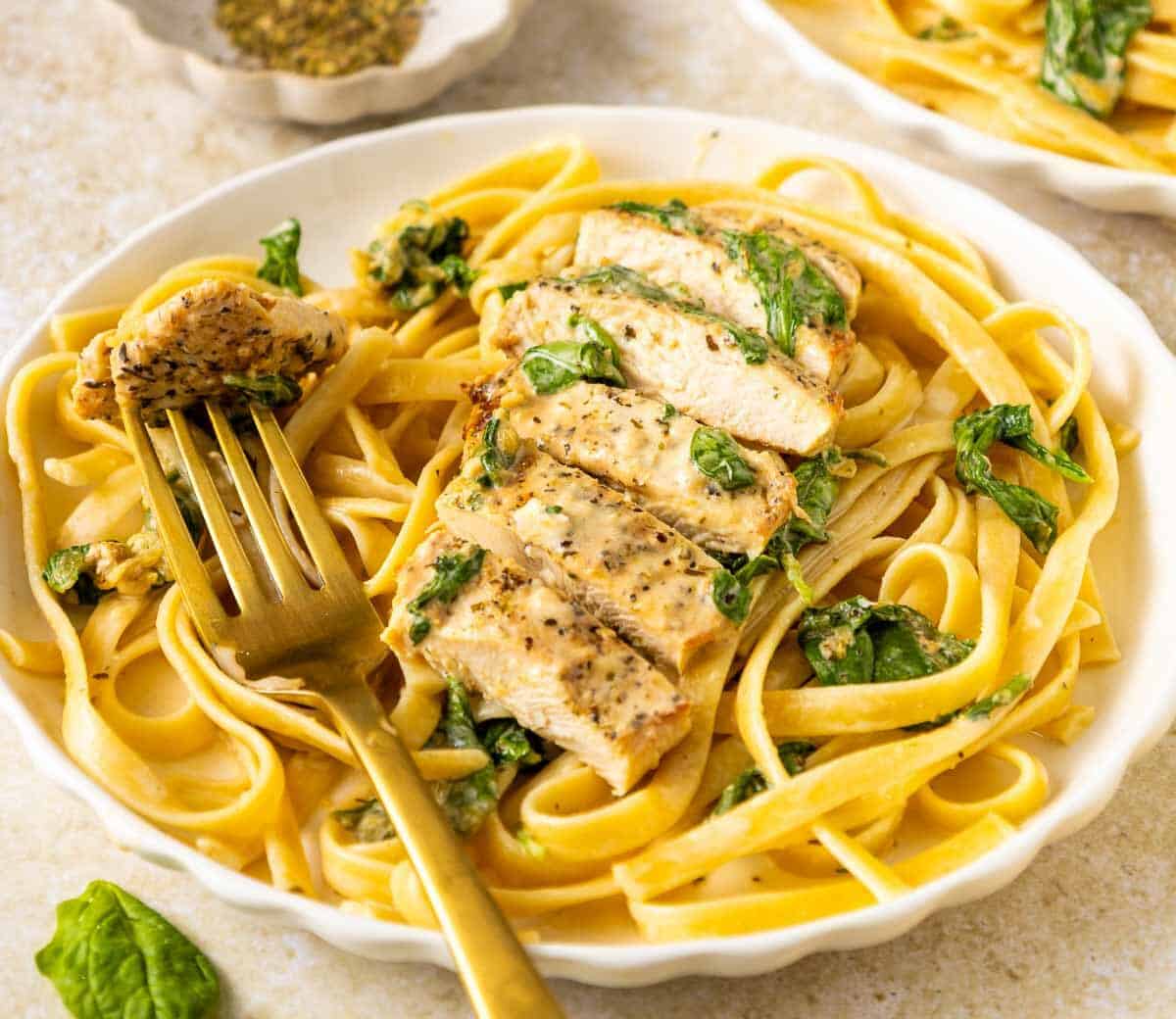 A plate of fettuccine pasta with spinach, topped with sliced grilled chicken. A golden fork holds a piece of chicken, and a small bowl of herbs is in the background.