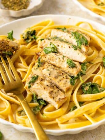 A plate of fettuccine pasta with spinach, topped with sliced grilled chicken. A golden fork holds a piece of chicken, and a small bowl of herbs is in the background.