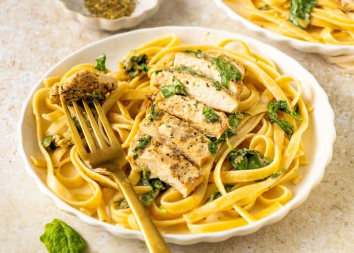 A plate of fettuccine pasta with spinach, topped with sliced grilled chicken. A golden fork holds a piece of chicken, and a small bowl of herbs is in the background.