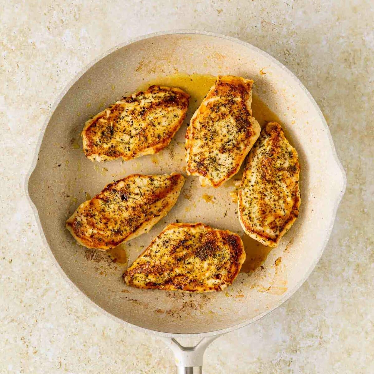 Five seasoned and cooked chicken breast pieces are arranged in a light-colored frying pan with a wooden handle, set on a textured, light beige surface.