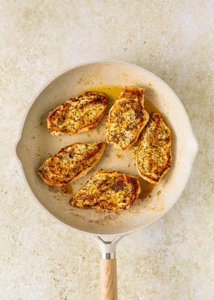 Five seasoned and cooked chicken breast pieces are arranged in a light-colored frying pan with a wooden handle, set on a textured, light beige surface.