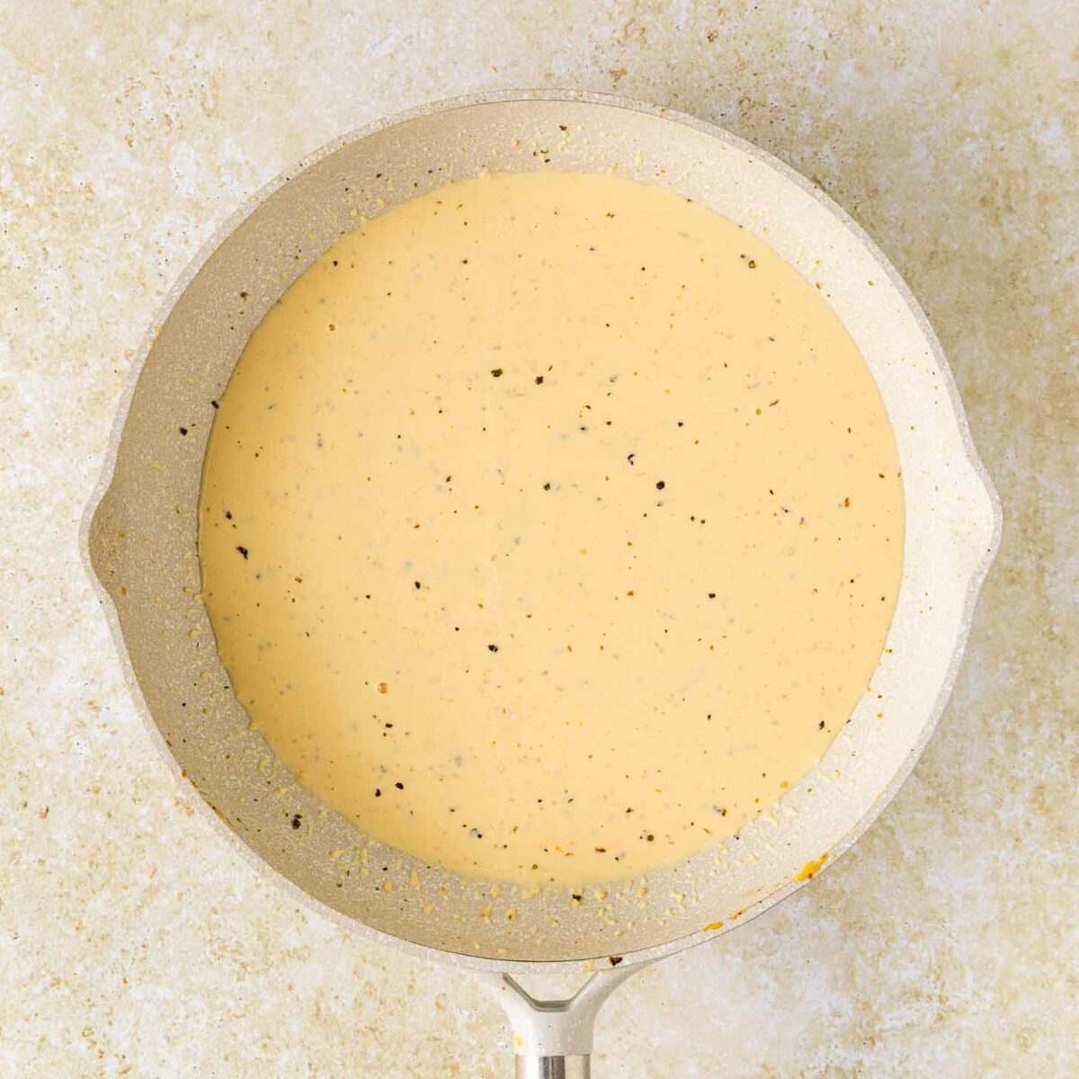 A beige frying pan with a wooden handle contains a creamy, light yellow sauce with visible black pepper specks, set on a light, textured surface.