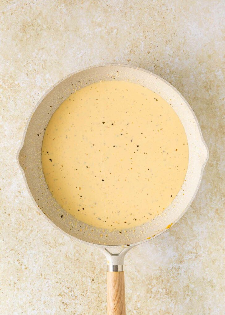 A beige frying pan with a wooden handle contains a creamy, light yellow sauce with visible black pepper specks, set on a light, textured surface.