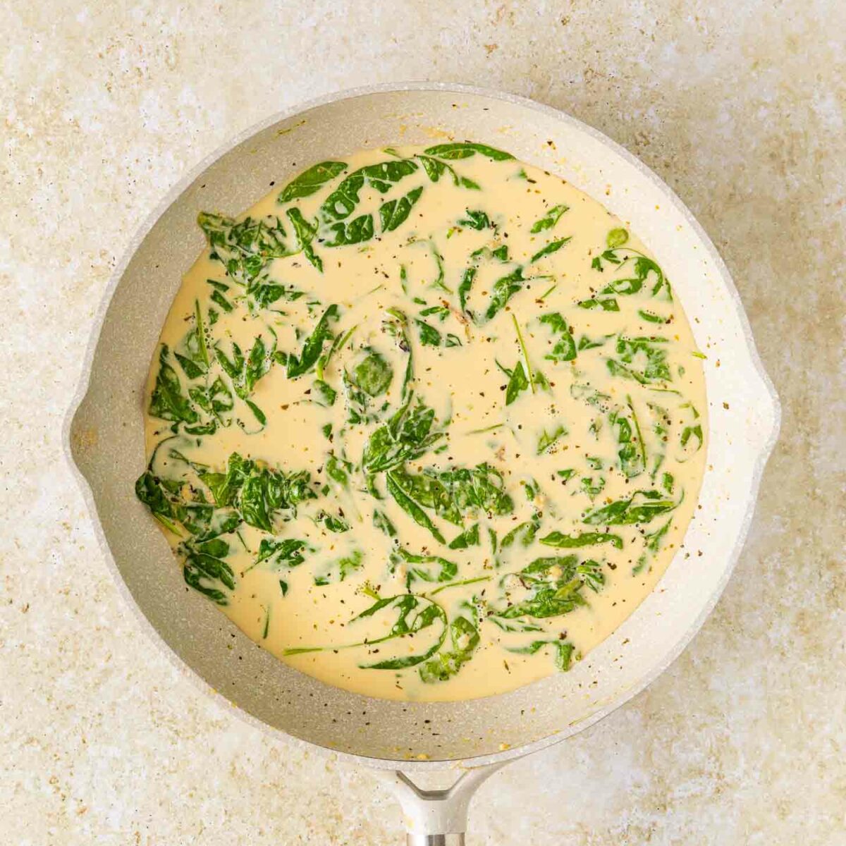 A light-colored skillet with creamy spinach sauce, featuring wilted spinach leaves in a pale yellow, seasoned cream, on a neutral countertop background.