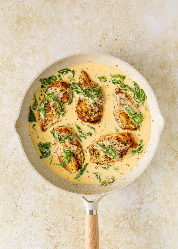 A skillet with four pieces of seasoned chicken breast in a creamy sauce, garnished with spinach leaves, sits on a light-colored surface.