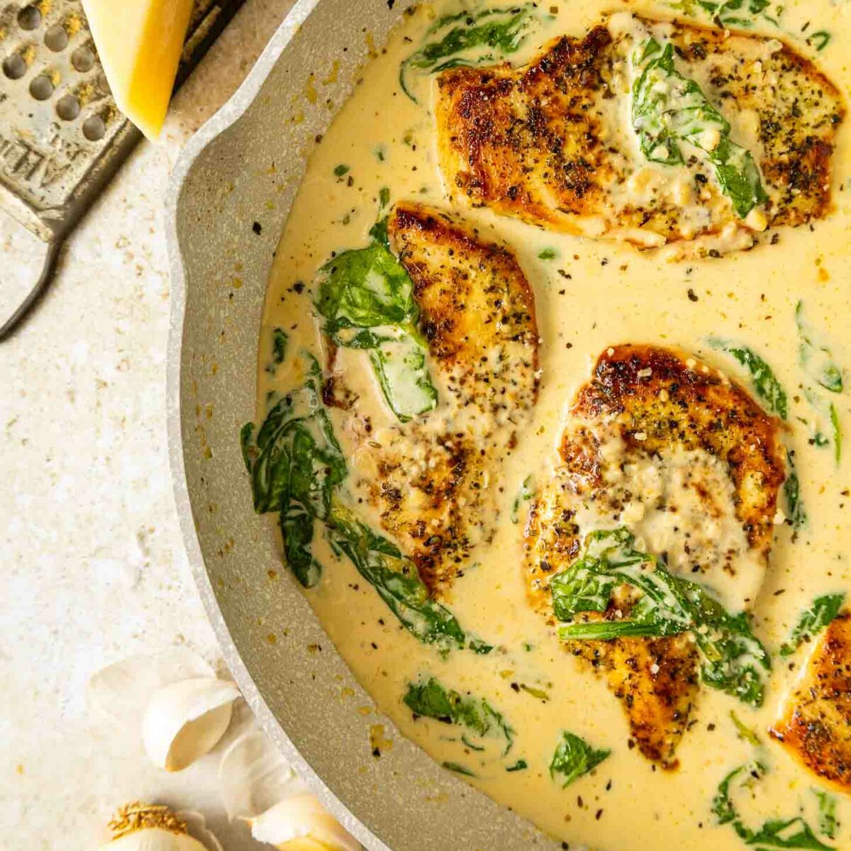 Creamy pan-seared chicken breasts in a skillet with spinach in a rich, golden sauce. A wedge of cheese, a grater, and garlic bulbs are visible nearby on a light surface.