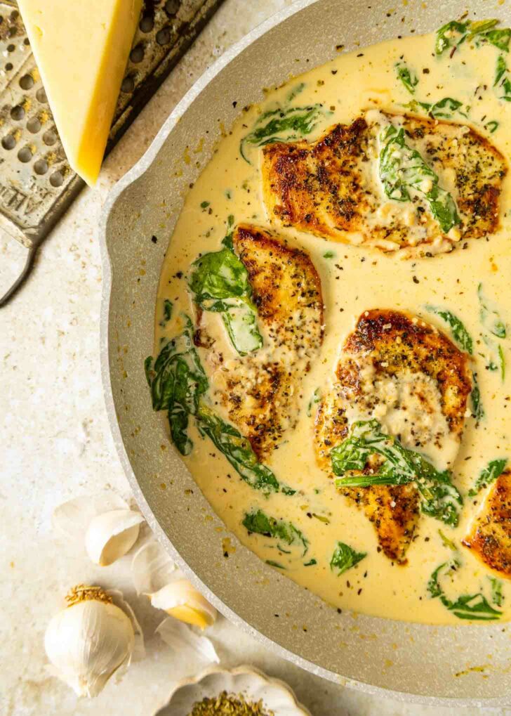 Creamy pan-seared chicken breasts in a skillet with spinach in a rich, golden sauce. A wedge of cheese, a grater, and garlic bulbs are visible nearby on a light surface.