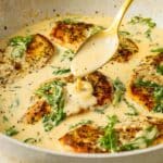 A skillet filled with golden-seared chicken breasts in a creamy sauce, garnished with wilted spinach. A hand holds a spoon, drizzling sauce over one of the chicken pieces.
