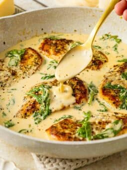 A skillet filled with golden-seared chicken breasts in a creamy sauce, garnished with wilted spinach. A hand holds a spoon, drizzling sauce over one of the chicken pieces.