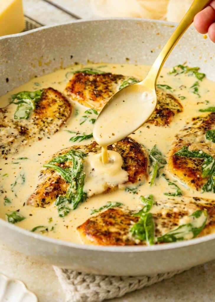 A skillet filled with golden-seared chicken breasts in a creamy sauce, garnished with wilted spinach. A hand holds a spoon, drizzling sauce over one of the chicken pieces.