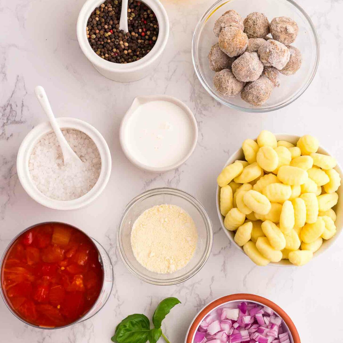A flat lay of ingredients on a marble surface, including gnocchi, chopped red onions, canned tomatoes, meatballs, heavy cream, salt, pepper, olive oil, garlic, and fresh basil leaves.