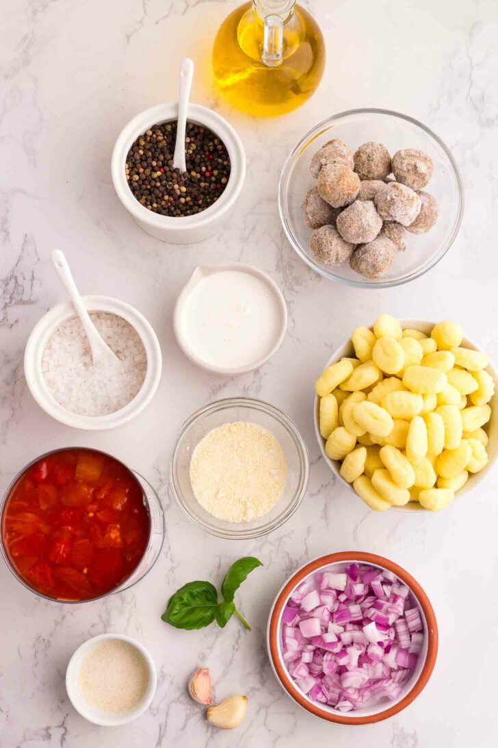 A flat lay of ingredients on a marble surface, including gnocchi, chopped red onions, canned tomatoes, meatballs, heavy cream, salt, pepper, olive oil, garlic, and fresh basil leaves.
