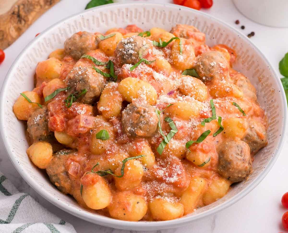 A bowl of gnocchi and meatballs in tomato cream sauce, garnished with grated cheese and fresh basil. Cherry tomatoes, basil leaves, and a striped napkin are nearby on a white surface.