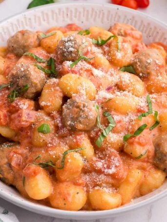 A bowl of gnocchi and meatballs in tomato cream sauce, garnished with grated cheese and fresh basil. Cherry tomatoes, basil leaves, and a striped napkin are nearby on a white surface.
