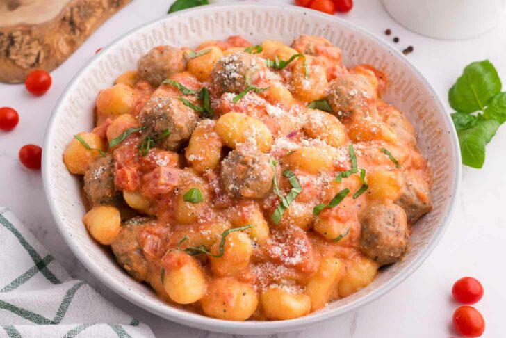 A bowl of gnocchi and meatballs in tomato cream sauce, garnished with grated cheese and fresh basil. Cherry tomatoes, basil leaves, and a striped napkin are nearby on a white surface.