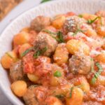 A bowl of gnocchi and meatballs in a creamy tomato sauce, garnished with fresh basil and grated Parmesan cheese.