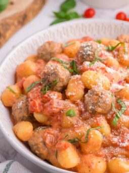 A bowl of gnocchi and meatballs in a creamy tomato sauce, garnished with fresh basil and grated Parmesan cheese.