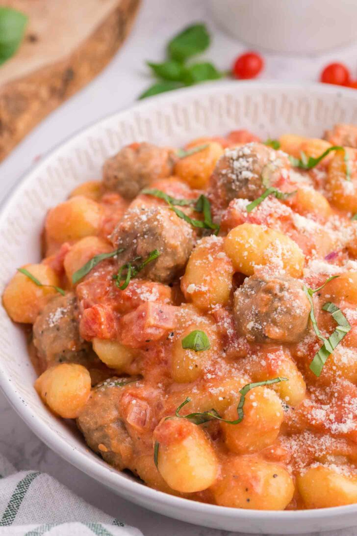 A bowl of gnocchi and meatballs in a creamy tomato sauce, garnished with fresh basil and grated Parmesan cheese.
