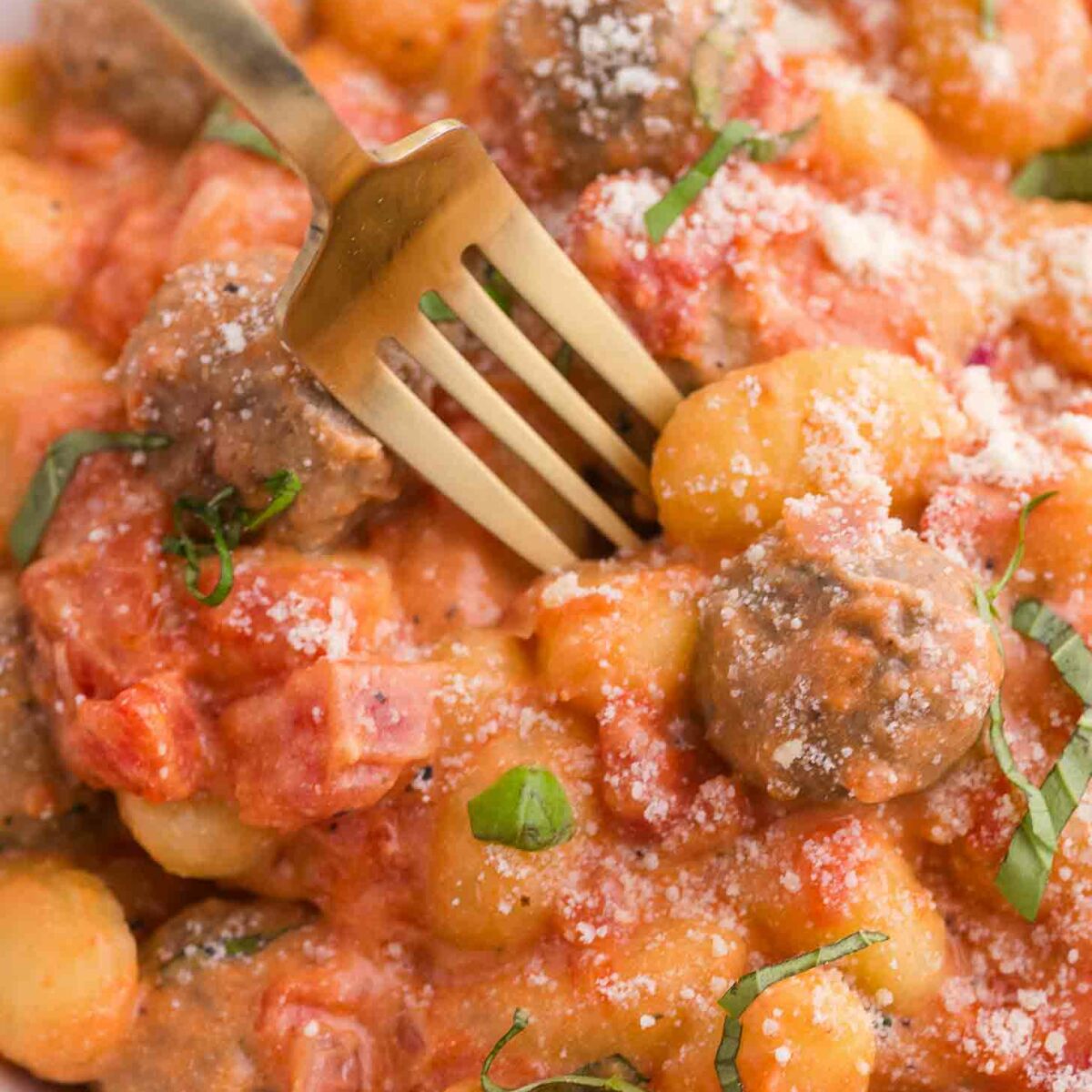 A close-up of gnocchi and meatballs in a creamy tomato sauce, garnished with grated cheese and fresh basil, with a gold fork resting on the dish.