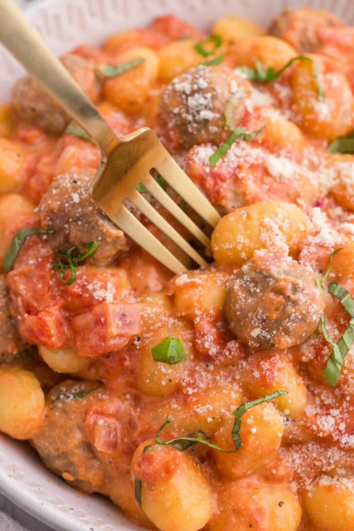 A close-up of gnocchi and meatballs in a creamy tomato sauce, garnished with grated cheese and fresh basil, with a gold fork resting on the dish.