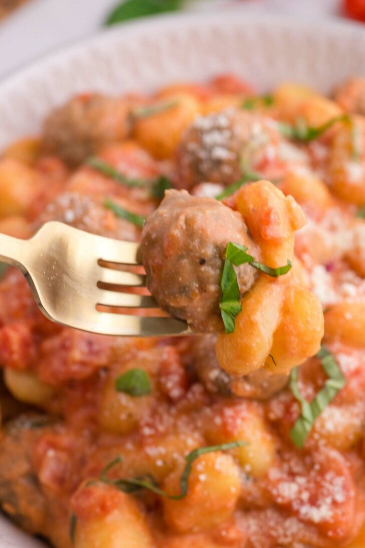 A fork holds up gnocchi and a meatball, both covered in tomato sauce, with fresh basil and grated cheese visible on top of a bowl of the same dish in the background.