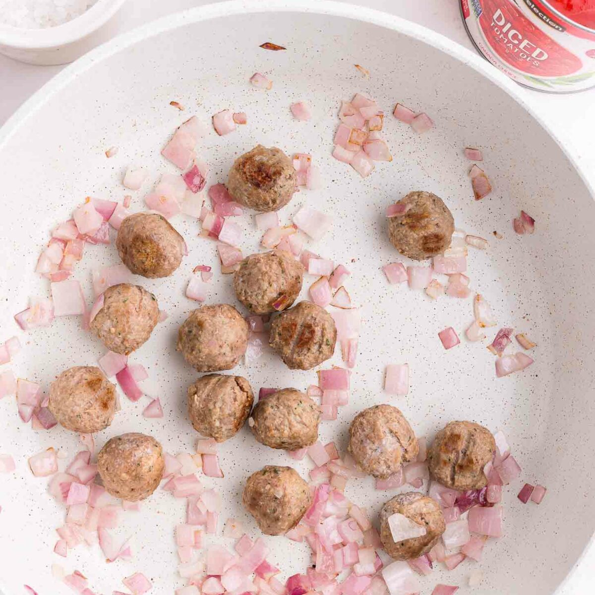 A white skillet with browned meatballs and chopped red onions being sautéed, with a can of diced tomatoes and a bowl of salt nearby on a white countertop.