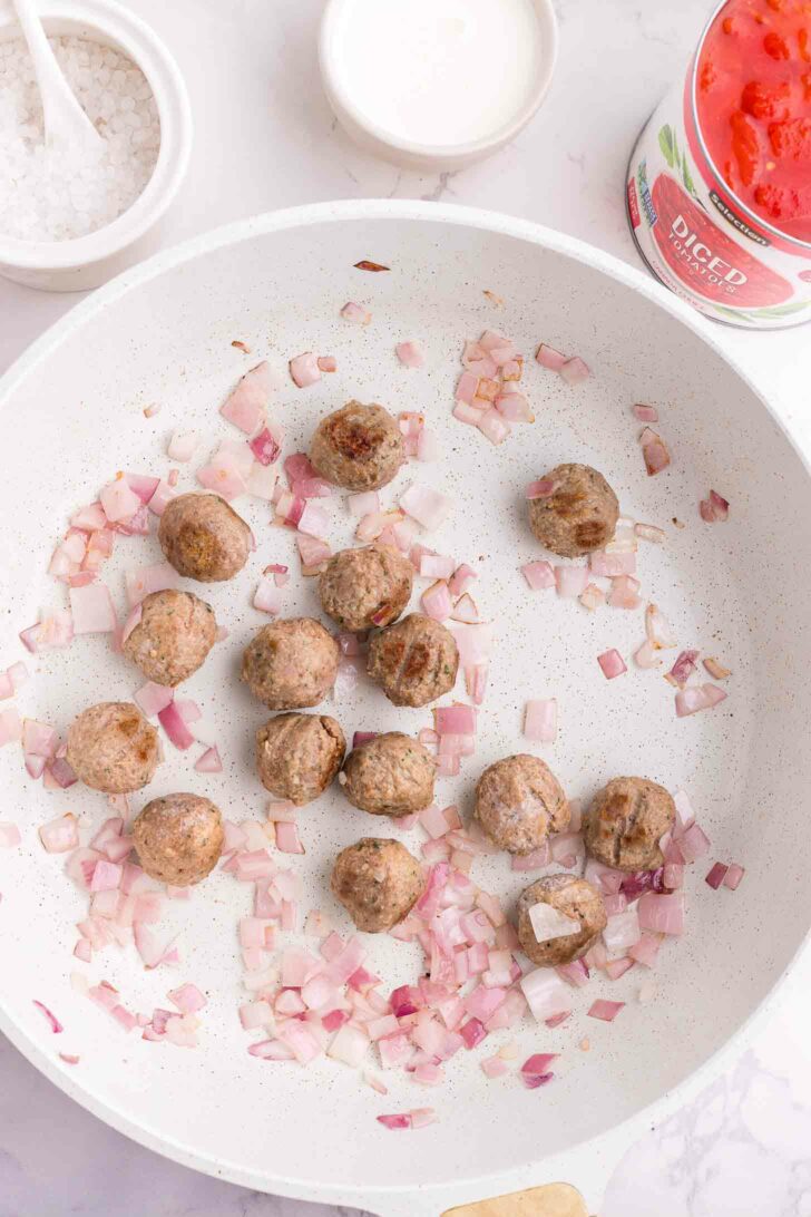 A white skillet with browned meatballs and chopped red onions being sautéed, with a can of diced tomatoes and a bowl of salt nearby on a white countertop.