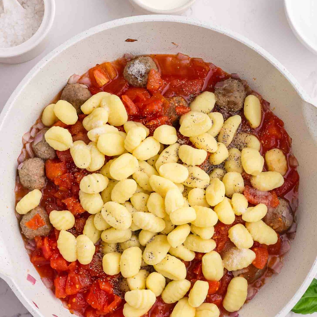 A white skillet filled with tomato sauce, meatballs, and uncooked gnocchi sits on a marble countertop, with small bowls of salt, cream, and pepper nearby.