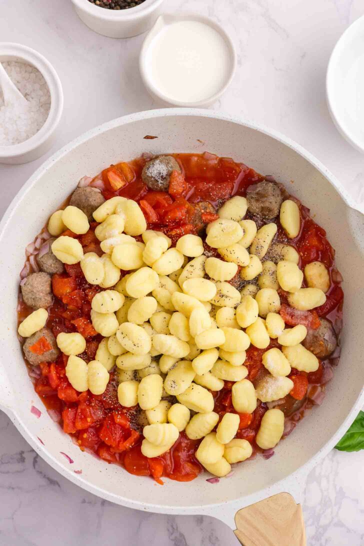 A white skillet filled with tomato sauce, meatballs, and uncooked gnocchi sits on a marble countertop, with small bowls of salt, cream, and pepper nearby.