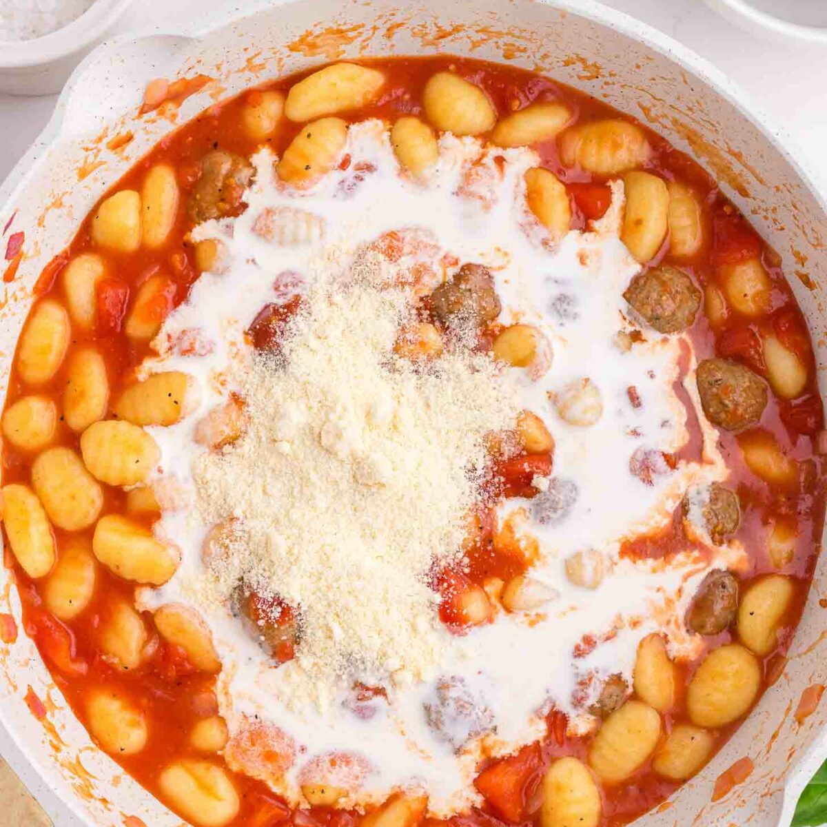 A skillet filled with gnocchi, meatballs, tomato sauce, cream, and grated parmesan cheese on top, ready to be mixed. The background shows a white countertop with a bowl of salt and a small plate.