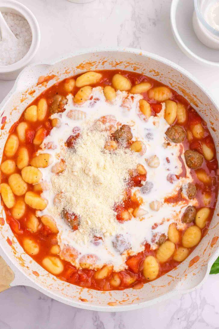 A skillet filled with gnocchi, meatballs, tomato sauce, cream, and grated parmesan cheese on top, ready to be mixed. The background shows a white countertop with a bowl of salt and a small plate.