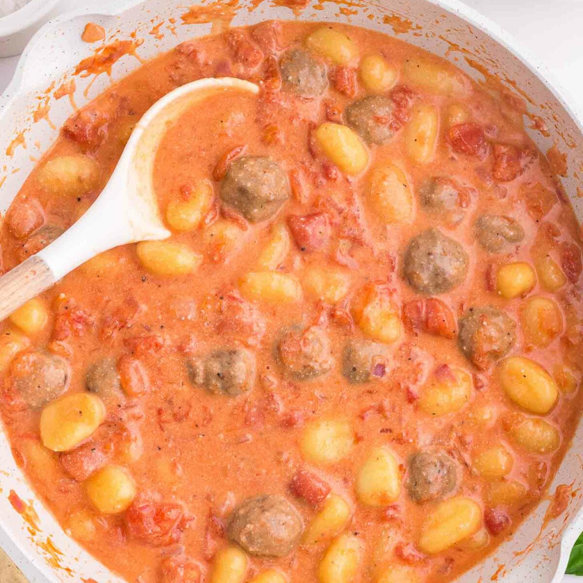 A white pan filled with gnocchi, meatballs, and diced tomatoes in a creamy tomato sauce, with a white spoon resting on the side. The pan sits on a marble countertop.