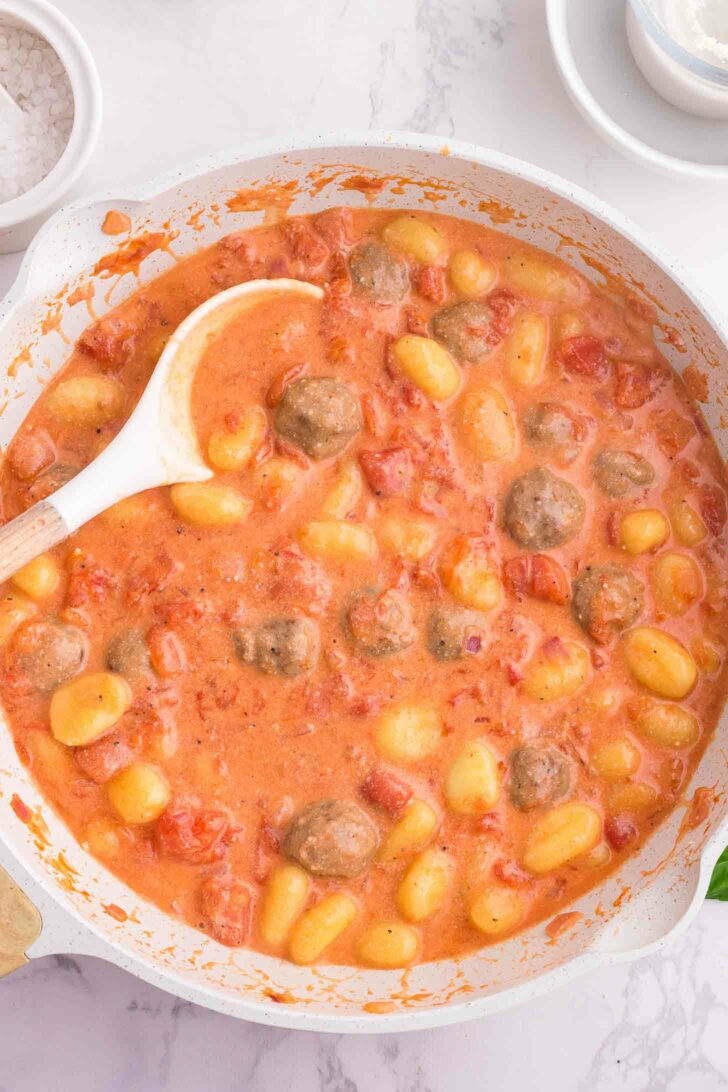 A white pan filled with gnocchi, meatballs, and diced tomatoes in a creamy tomato sauce, with a white spoon resting on the side. The pan sits on a marble countertop.
