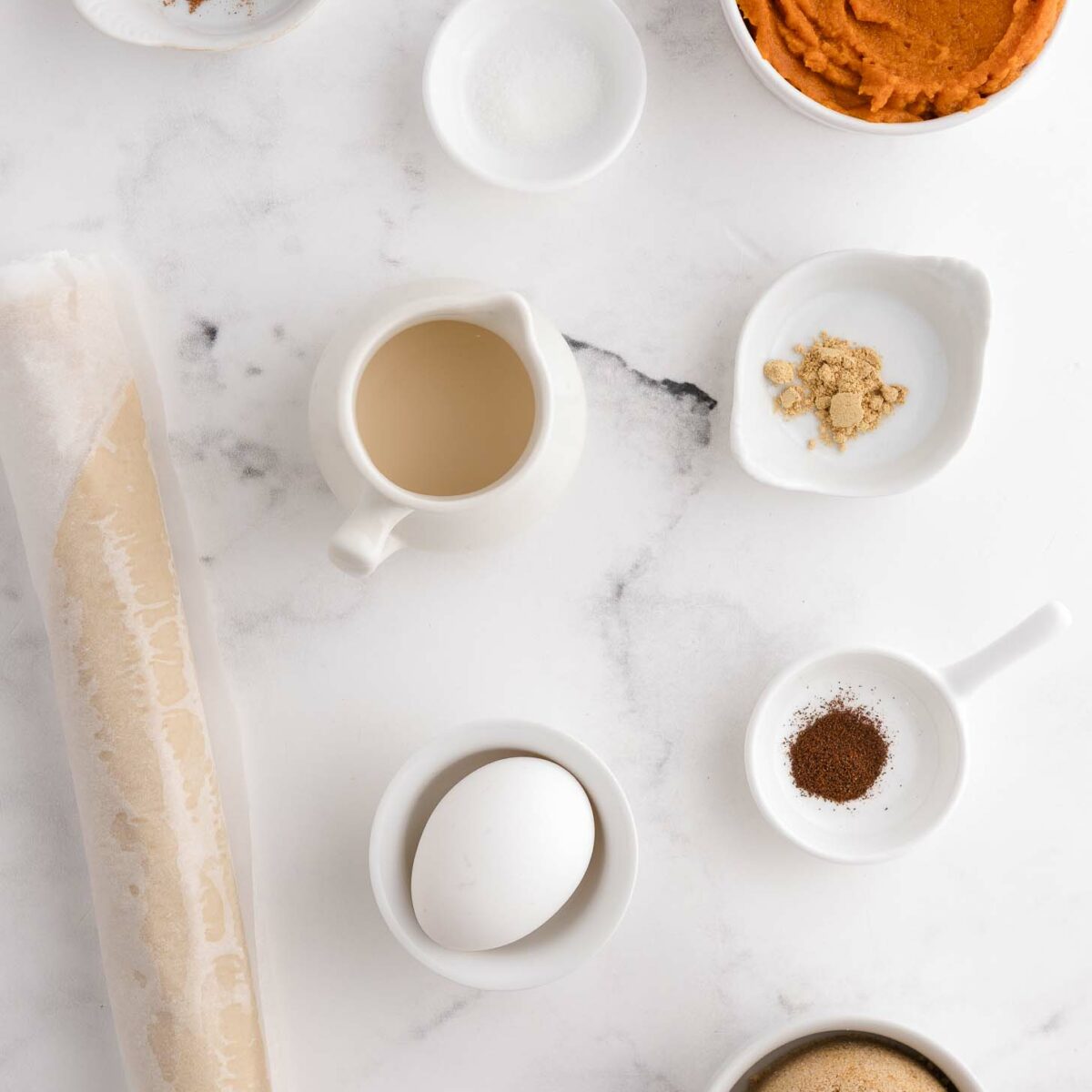 Baking ingredients on a white marble surface: pumpkin puree, brown sugar, egg, pie crust dough, milk, ground cinnamon, ginger, cloves, and salt in small bowls and containers.