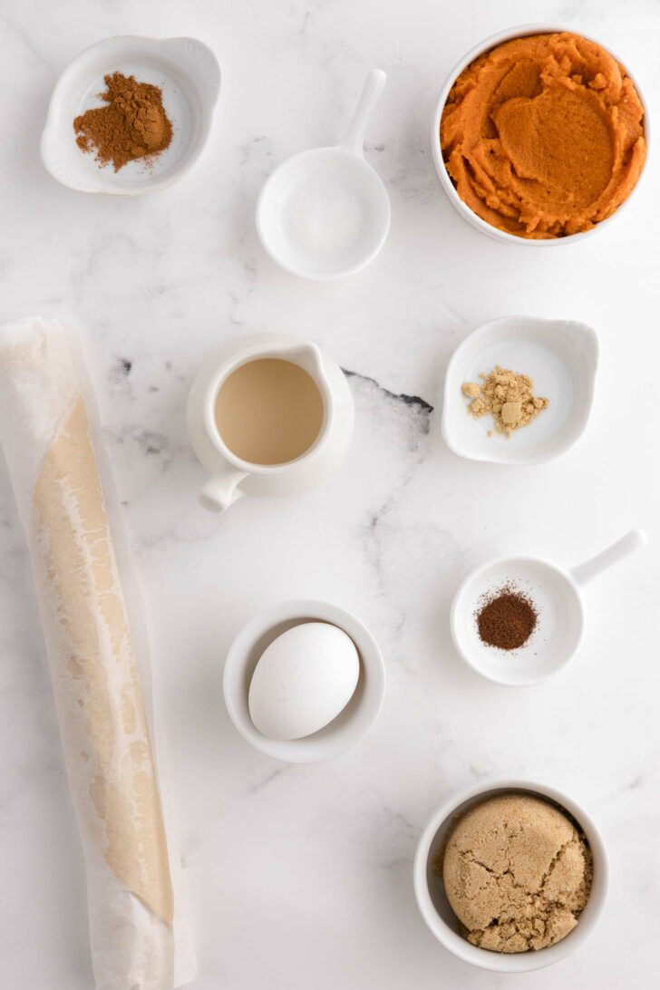 Baking ingredients on a white marble surface: pumpkin puree, brown sugar, egg, pie crust dough, milk, ground cinnamon, ginger, cloves, and salt in small bowls and containers.