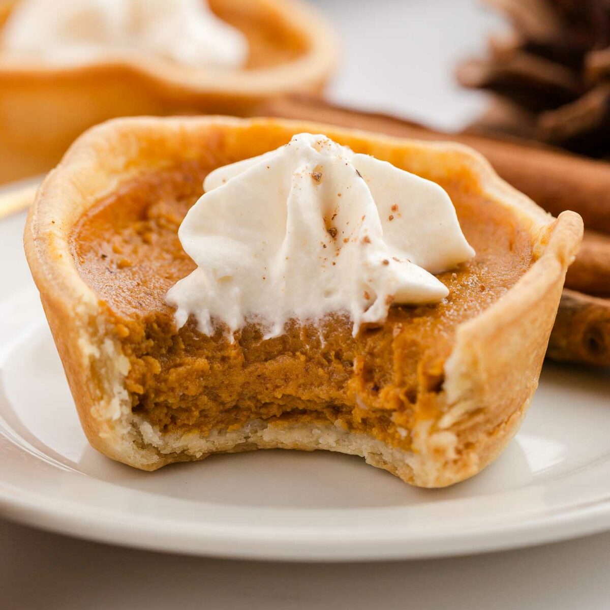A mini pumpkin pie with a bite taken out, topped with a swirl of whipped cream, sits on a white plate. Cinnamon sticks and another pie are blurred in the background.