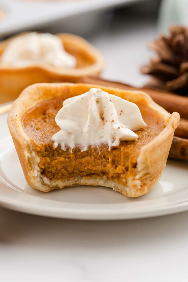 A mini pumpkin pie with a bite taken out, topped with a swirl of whipped cream, sits on a white plate. Cinnamon sticks and another pie are blurred in the background.