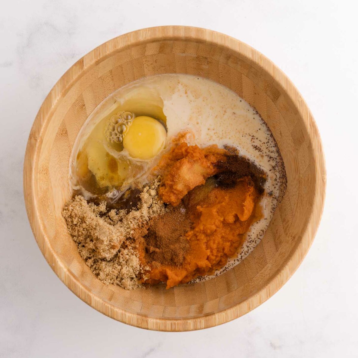 A wooden bowl filled with an egg, milk, brown sugar, pumpkin puree, and spices, all unmixed, on a white marble surface.