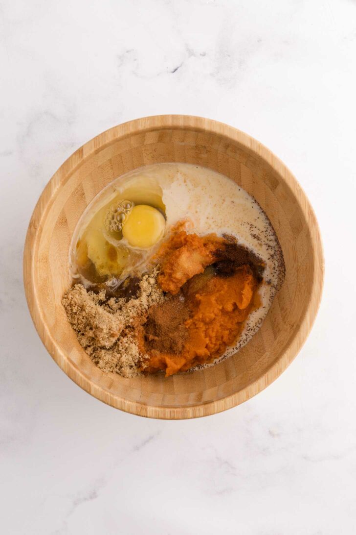 A wooden bowl filled with an egg, milk, brown sugar, pumpkin puree, and spices, all unmixed, on a white marble surface.