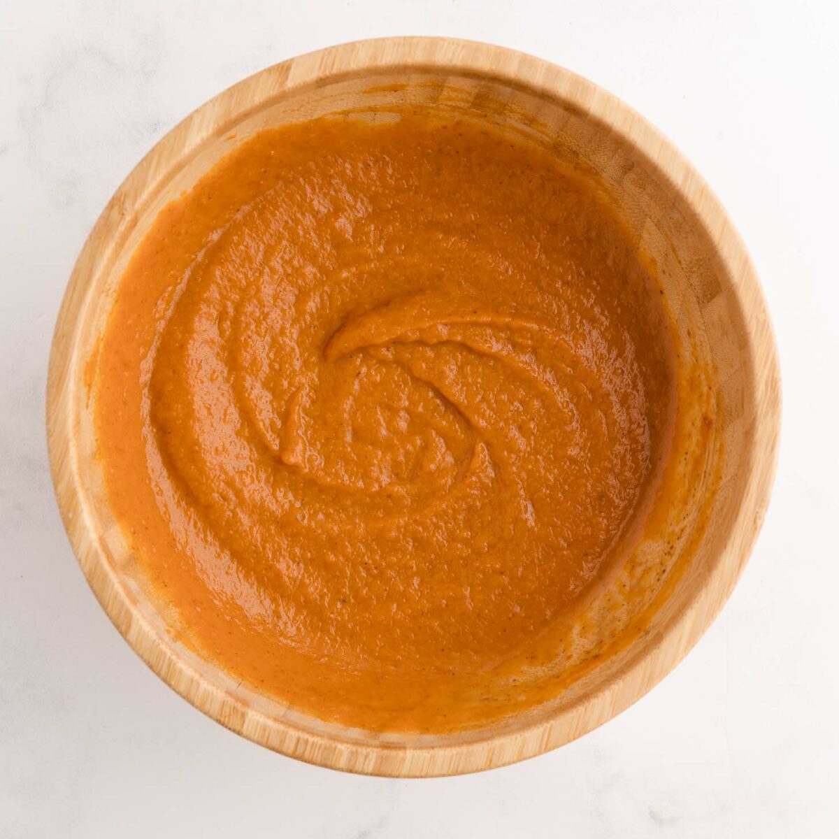 A wooden bowl filled with smooth, orange-pumpkin mixture, placed on a white marble surface.